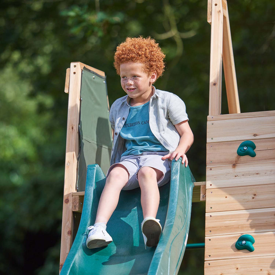 A young boy sitting on top of the wave slide of Plum Play's Climbing Pyramid Wooden Climbing Frame