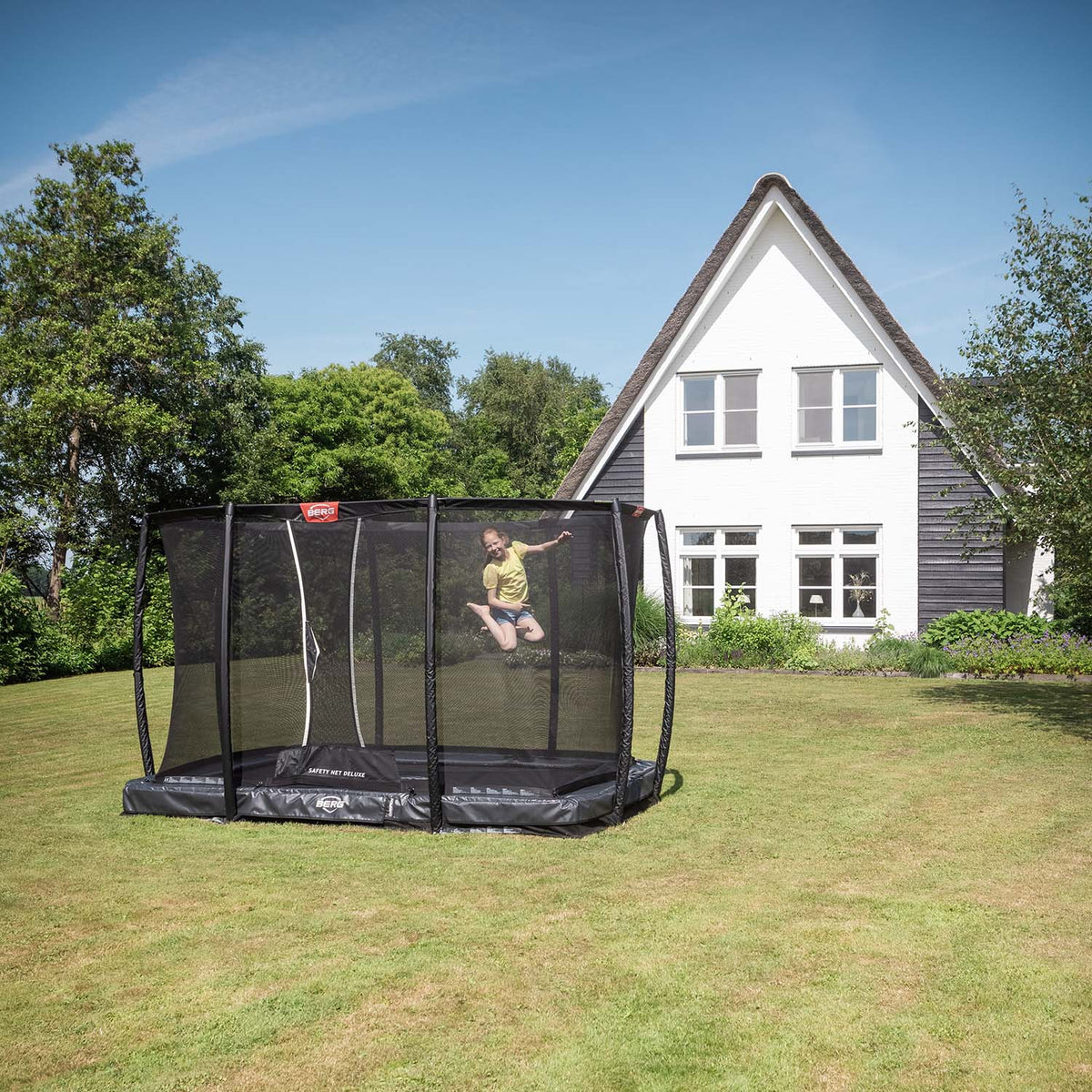 girl playing inside the BERG Ultim Champion InGround Rectangular Trampoline with Safety Net Deluxe in Grey colour in front of a white house