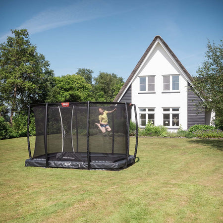 girl playing inside the BERG Ultim Champion InGround Rectangular Trampoline with Safety Net Deluxe in Grey colour in front of a white house